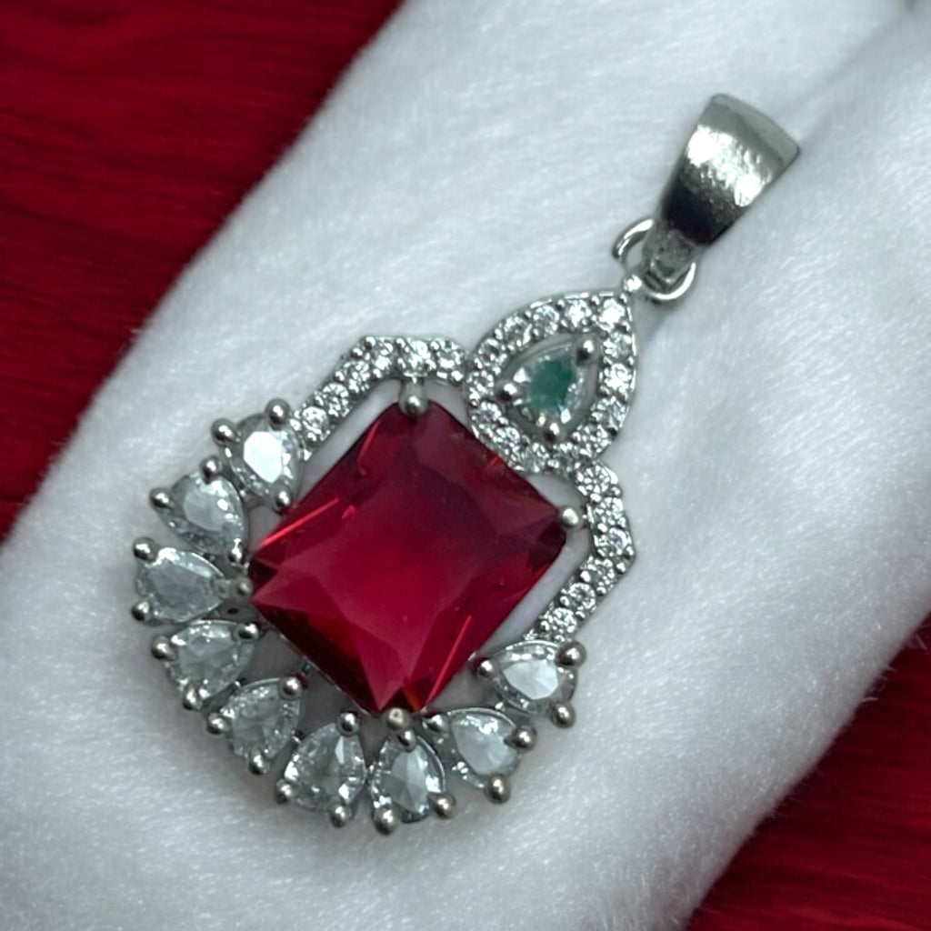 Silver-tone ruby-red pendant with white pear-cut halo and emerald accent, flat-lay close-up
