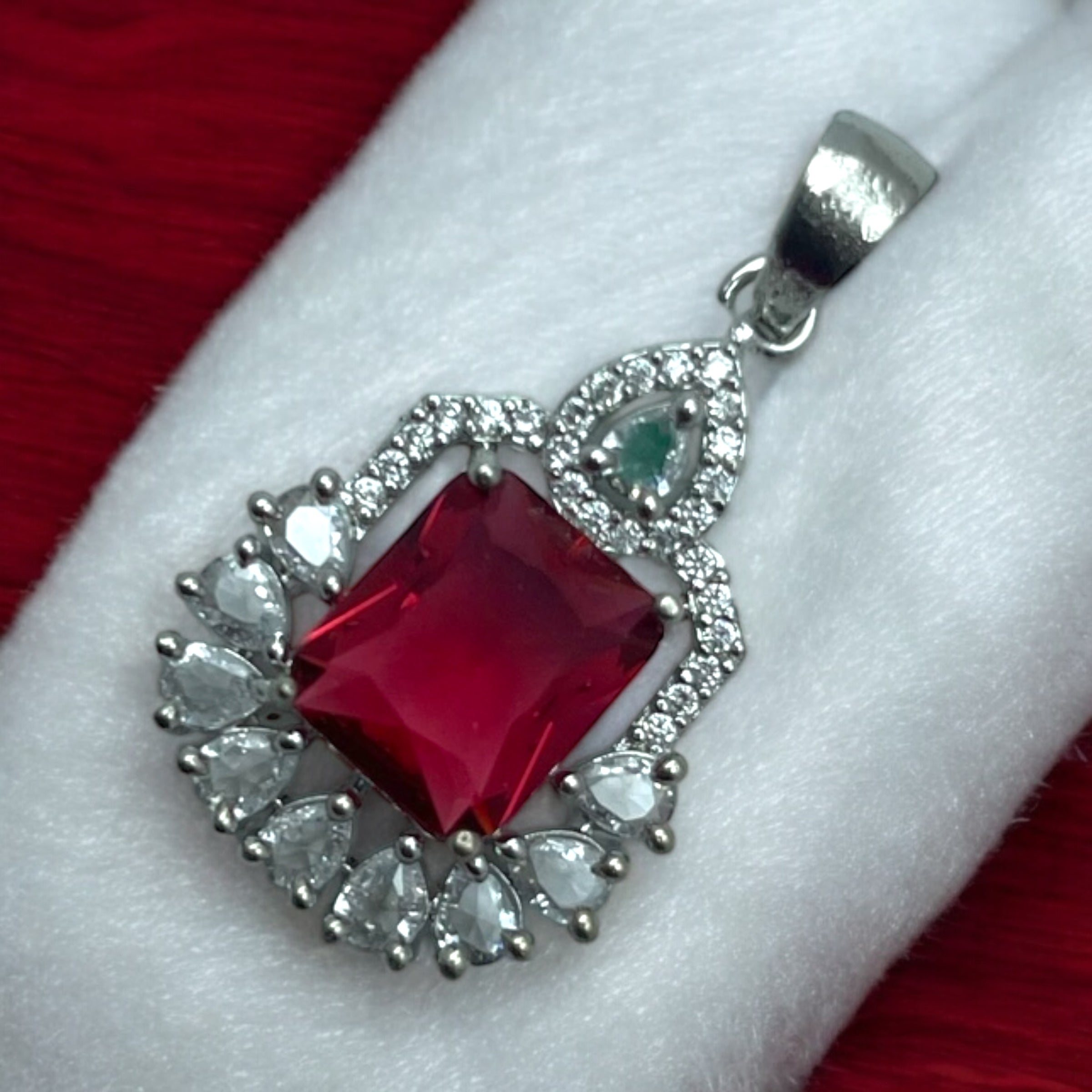 Silver-tone ruby-red pendant with white pear-cut halo and emerald accent, flat-lay close-up