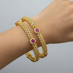 Gold-plated bangle set with crystal rows and oval ruby-red stones, worn stacked on wrist with water droplets