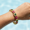 Gold-plated floral gemstone bangle with green and ruby CZ stones, worn on wrist with water droplets