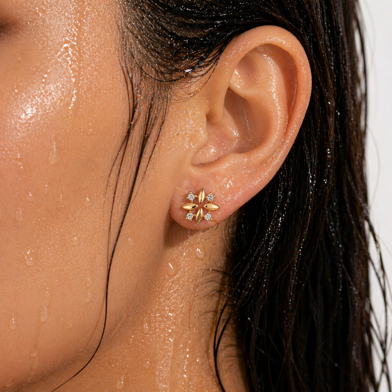 Close-up of an ear wearing a gold flower-shaped earring with small AD diamonds, against a blurred background.