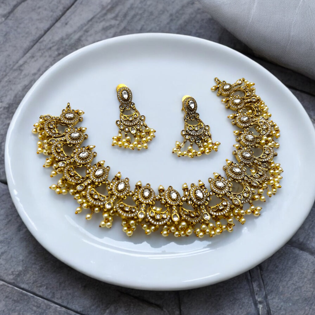 Antique gold Kundan necklace and earrings set with white stones and pearl fringe, flat-lay on a white plate
