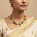 Woman wearing a gold necklace with green and red stones, pearl earrings, and a cream saree with gold patterns.