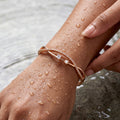 Rose gold crisscross bangle bracelet with crystal and pearl accents, worn on wrist near water