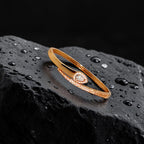 Rose gold-toned Aurelle bangle bracelet with pear-cut crystal centre and pave band, flat-lay on wet black stone