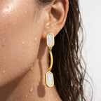 Gold-tone curved drop earrings with pearlescent oval inlays, worn on ear with water droplets showing waterproof finish