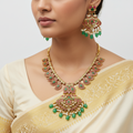 Woman wearing a gold necklace and earrings with green gemstones on a plain background