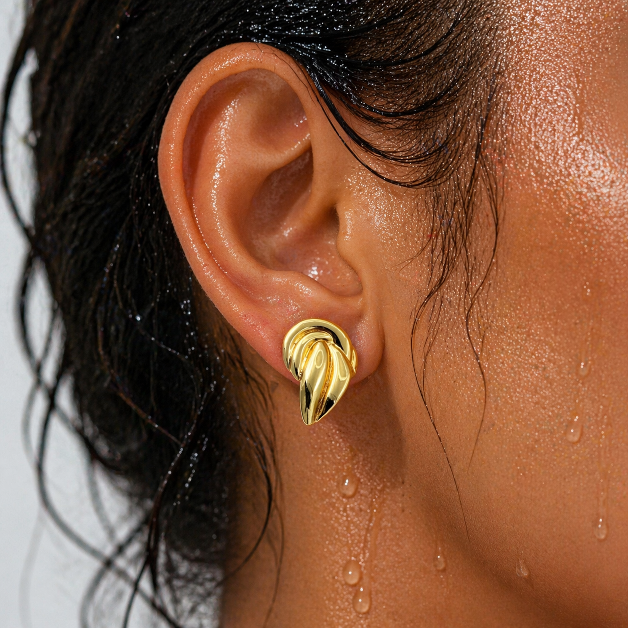 Close-up of an ear wearing a gold earring with water droplets on skin and hair.