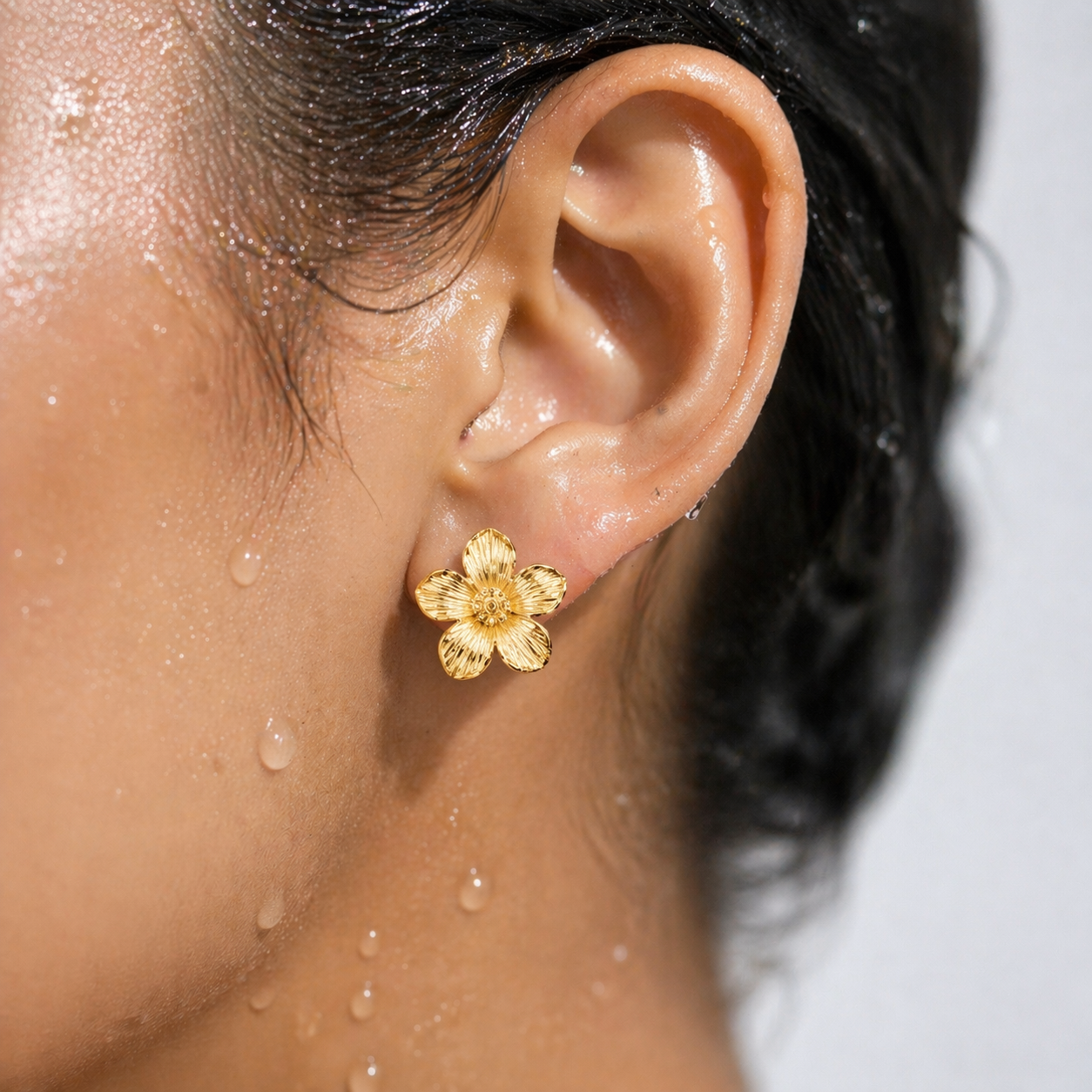 Close-up of an ear wearing a gold flower earring with water droplets on skin and hair.