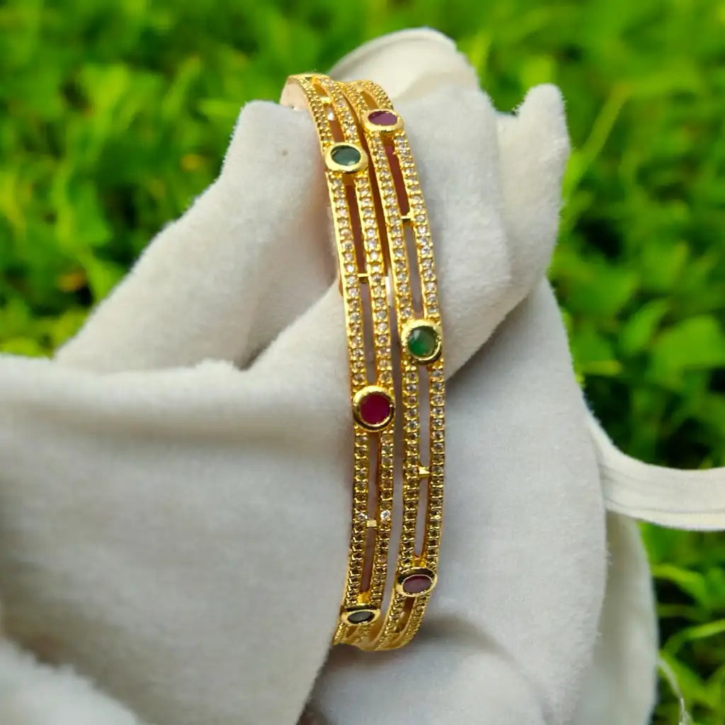 Gold-plated gemstone bangle set with emerald and ruby stones and pave crystals, worn stacked on wrist with water droplets