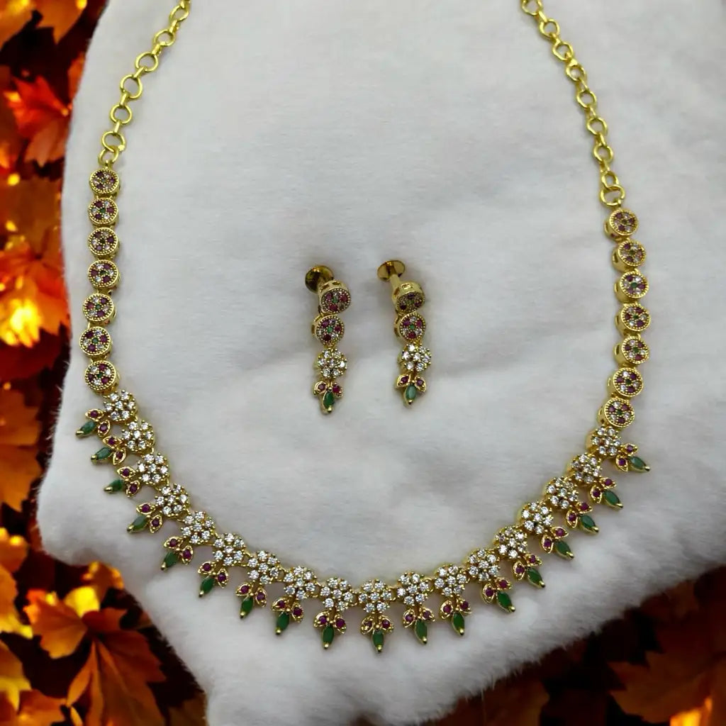 Gold-plated floral crystal necklace and drop earrings set with ruby and green accents, flat-lay on white fabric