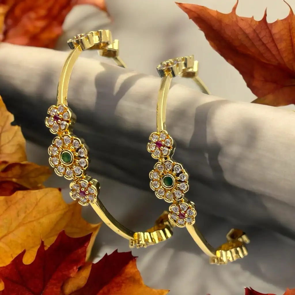 Gold floral crystal bangles with emerald-green centres, pair displayed upright against autumn leaves