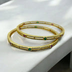 Gold-plated crystal-studded bangle pair with emerald and ruby stones, flat-lay close-up on marble