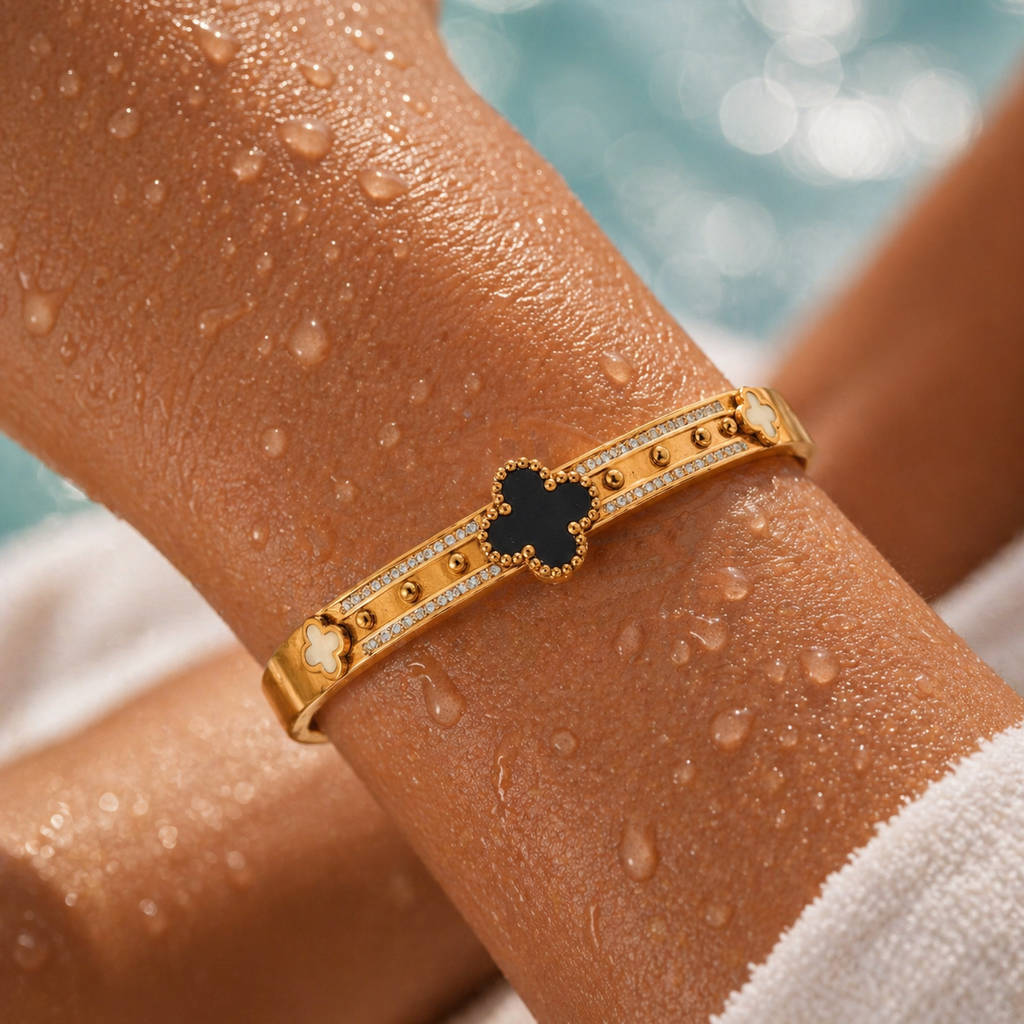 Gold bracelet with a black clover charm on a wet arm by a pool.