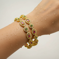 Gold-plated marquise bangle set with ruby and emerald-tone stones in crystal halos, worn stacked on wrist with water droplets